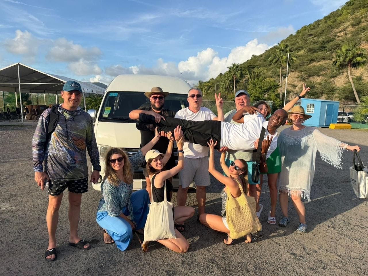 A joyful group of guests lifting Andy in the air beside the white Toyota HiAce at sunset in Sint Maarten